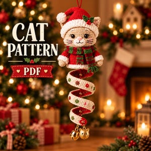 Op de afbeelding: Een gehaakte kattenhanger met een rode kerstmuts en een rood-groen gestreepte sjaal. Onder de kat hangt een lang, spiraalvormig, crèmekleurig lint versierd met kleine groene en rode bloemmotieven, eindigend in twee goudkleurige kattenbelletjes.