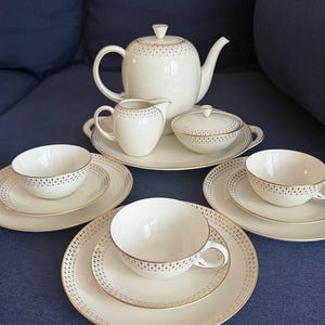 Puede incluir: Un juego de té de porcelana blanca con detalles dorados. El juego incluye una tetera, una jarra de crema, una azucarera y varias tazas y platillos. El juego de té tiene un delicado patrón de pequeños puntos dorados y un borde dorado. El juego está dispuesto sobre una superficie azul.