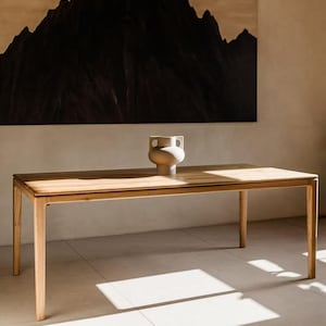 Op de afbeelding: Een rechthoekige eettafel van lichtgekleurd hout met vier rechte poten en een glad oppervlak. In het midden van de tafel staat een matte, gebroken witte keramische vaas met een ronde vorm en twee kleine, platte handvatten aan de hals.