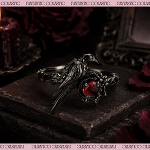 May include: A dark metallic bracelet featuring a sculpted bird perched next to a deep red circular gemstone. The band has a textured, organic design that wraps around the wrist, showcasing intricate detailing on the bird's feathers and the setting.