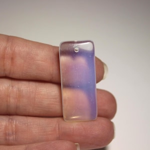 May include: A rectangular pendant with a hole at the top. The pendant has a translucent appearance with a gradient of colors, including pink, purple, and white. The pendant is held in a hand.