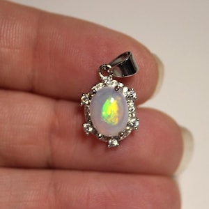 May include: A silver-toned pendant featuring a central oval-shaped iridescent opal gemstone with flashes of green and yellow. The opal is surrounded by a halo of small, clear, round-cut stones set in a metal frame with a top bail for a chain.