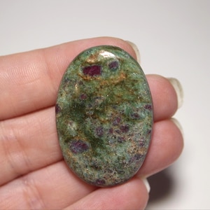 May include: An oval-shaped polished gemstone featuring a mottled green base color with embedded deep red and magenta inclusions throughout the surface. The stone has a smooth, glossy finish and displays natural variations in texture and color.