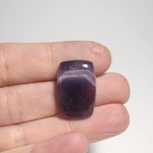 May include: A polished, rectangular amethyst gemstone with a deep purple hue, accented by bands of white and lighter purple. The stone is held in a hand, showcasing its smooth, rounded edges and natural color variations.