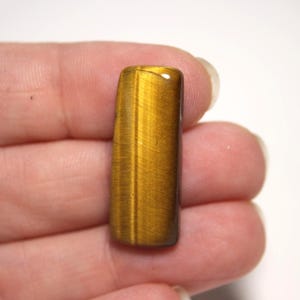 May include: A rectangular tiger's eye gemstone cabochon featuring a smooth, polished surface with a distinct chatoyant golden-brown luster and vertical bands of light and dark brown tones throughout the stone's natural crystalline structure.