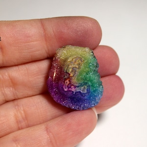 Puede incluir: Un cabujón de ágata arcoíris con una variedad de colores, incluyendo rojo, naranja, amarillo, verde, azul y morado. La ágata es translúcida y tiene una textura cristalina.