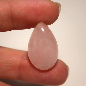 May include: A smooth, polished rose quartz gemstone shaped like a teardrop. The stone features a translucent, pale pink color with subtle internal cloudiness and a reflective surface that catches the light, showcasing its natural, rounded, and symmetrical form.