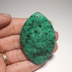 May include: A green gemstone pendant with black speckles, teardrop-shaped with a drilled hole at the top. The pendant is held in a hand, showcasing its polished surface and natural color variations. A gold ring is visible on the hand.
