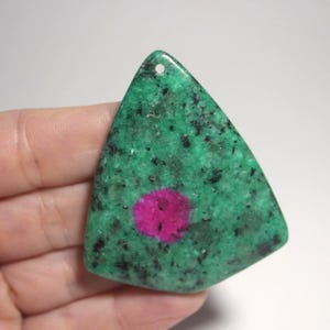 May include: A triangular-shaped gemstone pendant with a hole at the top. The stone features a speckled green base with a vibrant pink center. The pendant is held in a hand.