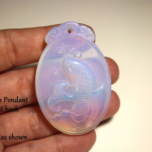 May include: A translucent, oval-shaped pendant with a carved koi fish design. The pendant is a blend of white, lavender, and pale blue hues. The fish is depicted swimming with bubbles, and the top has a decorative loop for a chain.