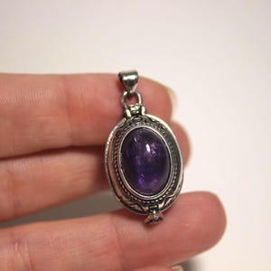 May include: Oval silver pendant with a large, polished purple amethyst stone. The pendant has an ornate, detailed frame and a small loop at the top for a chain. The amethyst is a deep, rich purple color.