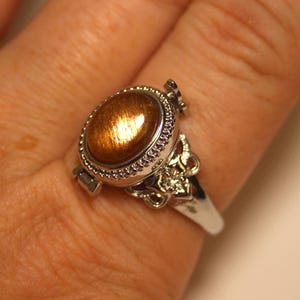 May include: A silver-toned ring featuring an oval-shaped, shimmering brown sunstone cabochon set within a beaded metal border. The band displays intricate metalwork details on the sides, including a small, clear, faceted stone accent.