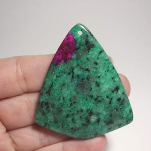May include: A triangular-shaped gemstone pendant with a hole at the top. The pendant features a vibrant green base with black speckles and a patch of deep pink. The stone is held in a hand.