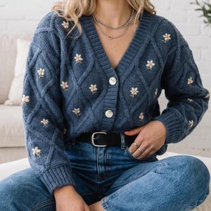 Puede incluir: Un cárdigan azul de punto trenzado con cuello en V, decorado con flores blancas bordadas con centros amarillos. La prenda cuenta con un cierre frontal de botones con dos botones blancos redondos y un patrón de rombos en toda la superficie.