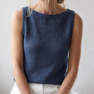 Puede incluir: Un top sin mangas de punto azul con cuello barco. El top está hecho de un material suave y texturizado. La modelo lleva pantalones blancos. El top es de color liso y parece ser una prenda cómoda.