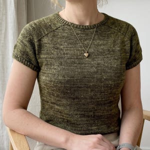 May include: A short-sleeved, crew-neck knit top in a variegated olive green yarn with a subtle heathered texture. The garment features raglan sleeves and a ribbed neckline, cuffs, and hem, paired with a simple gold-toned heart pendant necklace.