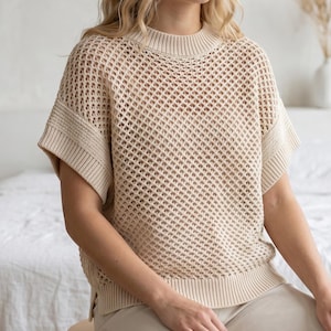 Puede incluir: Un suéter beige de manga corta que presenta un patrón de malla de rombos calados en todo el cuerpo. La prenda incluye un cuello redondo acanalado, puños acanalados en las mangas y un dobladillo ancho acanalado, creando una silueta estructurada.