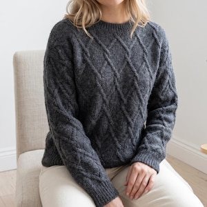 May include: A dark charcoal gray long-sleeve sweater featuring a prominent diamond-shaped cable knit pattern across the front and sleeves. The garment has a classic crew neckline and ribbed cuffs, providing a textured look with a consistent knit.