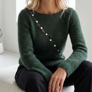 May include: A dark forest green long-sleeved sweater featuring a diagonal opening across the chest. The opening is secured with a row of small, round, white buttons that extend from the neckline down toward the side of the waist.