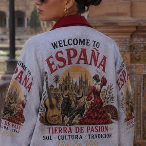 Könnte beinhalten: Graue Jacke mit rotem Kragen und Bündchen, mit dem Text "WELCOME TO ESPAÑA". Auf der Rückseite sind eine Gitarre, ein Stier, eine Flamenco-Tänzerin und die Worte "TIERRA DE PASION" abgebildet.
