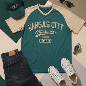 Może przedstawiać: Codzienny str&oacute;j składający się z zielono-beżowej koszulki graficznej Kansas City Missouri, pasującej czapki z daszkiem, ciemnych szort&oacute;w dżinsowych, białych trampek, okular&oacute;w przeciwsłonecznych i srebrnego zegarka. Koszulka ma napis "KANSAS CITY Missouri EST. 1838 KCMO".