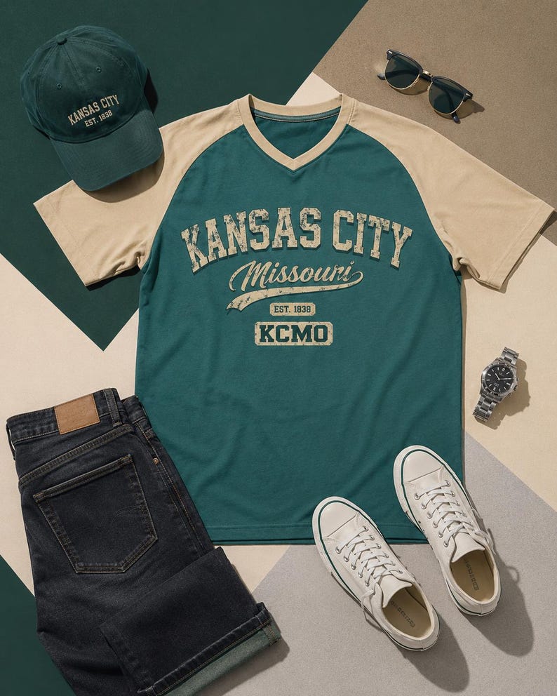 Może przedstawiać: Codzienny str&oacute;j składający się z zielono-beżowej koszulki graficznej Kansas City Missouri, pasującej czapki z daszkiem, ciemnych szort&oacute;w dżinsowych, białych trampek, okular&oacute;w przeciwsłonecznych i srebrnego zegarka. Koszulka ma napis "KANSAS CITY Missouri EST. 1838 KCMO".