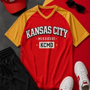 Può includere: Un completo a tema Kansas City rosso e giallo, che include un berretto da baseball, una maglietta, pantaloncini e scarpe da ginnastica. La maglietta presenta la scritta "Kansas City Missouri KCMO". Le scarpe da ginnastica sono bianche con dettagli rossi. I pantaloncini sono neri.