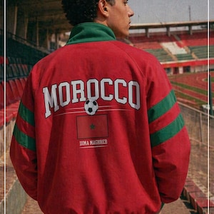 Può includere: Giacca rossa con dettagli verdi e la scritta "MOROCCO" in bianco. Un pallone da calcio e la bandiera marocchina sono sotto. Il testo "DIMA MAGHREB" è sotto la bandiera. La giacca ha strisce verdi sulle maniche.