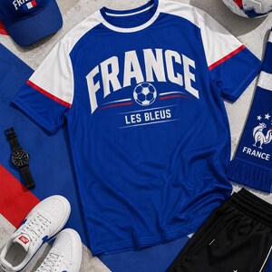 Puede incluir: Un conjunto de art&iacute;culos con tem&aacute;tica francesa: una camiseta azul con "FRANCE" y un bal&oacute;n de f&uacute;tbol, una gorra a juego, una bufanda, pantalones cortos negros, un bal&oacute;n de f&uacute;tbol, un reloj y zapatillas blancas con detalles azules. El texto "LES BLEUS" tambi&eacute;n es visible.