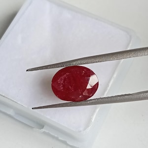 May include: An oval-cut, deep red ruby gemstone held by a pair of metal tweezers. The stone features multiple facets that reflect light, showcasing a rich, saturated crimson colour and a polished, translucent appearance with visible internal inclusions.