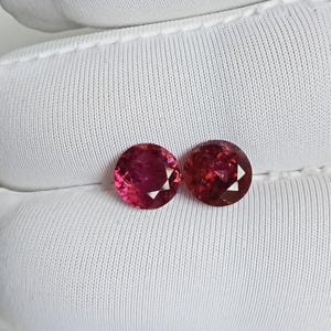 May include: Two round, faceted, deep pink gemstones with a brilliant cut. The stones exhibit a vibrant, saturated color with multiple triangular facets that reflect light, creating a sparkling appearance against a white fabric background.