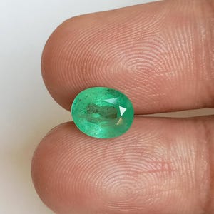 Op de afbeelding: Een ovaalvormige, gefacetteerde groene edelsteen met zichtbare interne insluitingen en een doorschijnend uiterlijk. De steen vertoont een levendige, verzadigde groene tint en is geslepen met meerdere platte oppervlakken die het licht reflecteren.