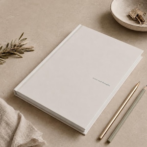May include: A cream-colored notebook with the words "notes and thoughts" printed on the cover. Two pencils, one gold and one gray, are positioned near the notebook. A small bowl with office supplies and a sprig of greenery are also in the image.