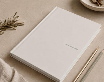 May include: A cream-coloured notebook with the words "notes and thoughts" printed on the cover. Two pencils, one gold and one grey, are positioned near the notebook. A small bowl with office supplies and a sprig of greenery are also in the image.