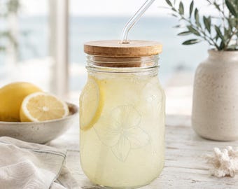 May include: A clear glass jar with a bamboo lid and a glass straw, filled with a yellow beverage and a lemon slice. The jar has a floral design etched on the side. A bowl of lemons and a vase are in the background.