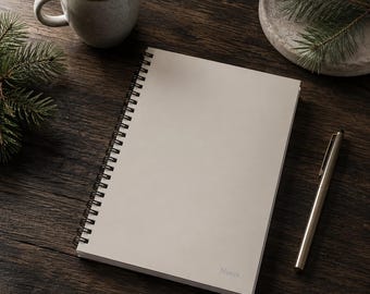May include: A spiral-bound notebook with a blank, off-white cover, and the word "Notes" printed in the bottom right corner. A silver pen rests beside the notebook on a dark wooden surface, with a cup of coffee and a small decorative dish with greenery.