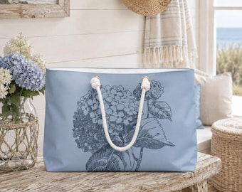 May include: A light blue tote bag featuring a dark blue floral illustration of hydrangeas. The bag has white rope handles and is made of a sturdy material. The tote bag is sitting on a rustic wooden table.