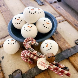 May include: A blue bowl filled with white felt snowman ornaments. The bowl is sitting on a wooden surface with a brown leather strap. There are red and white striped candy canes and a tag that says "Happy Holidays" on the surface.