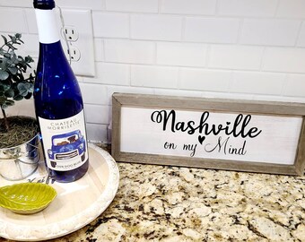 May include: A wooden sign with the text "Nashville on my ♥ Mind" in black script font. The sign is framed in a light brown wood frame and is sitting on a granite countertop.