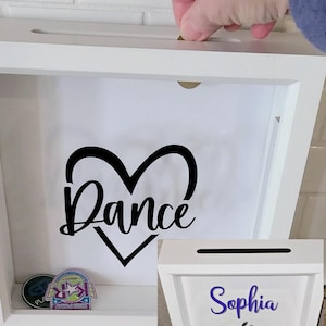 May include: White shadow box with a slot for inserting items. The box features a black heart with the word "Dance" inside. A hand is inserting a coin into the slot. Another box has the name "Sophia" and a dancer silhouette.