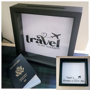 May include: Black shadow box with a white background and the text "travel is my therapy" with a plane and heart graphic. A US passport is in the foreground.