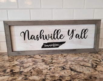 May include: A framed wooden sign with the text "Nashville Y'all" in black script font. The sign also features a black outline of the state of Tennessee with the word "Tennessee" written inside.