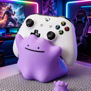 May include: A white game controller rests in a purple, amorphous, blob-shaped stand with two small black dots for eyes and a thin black line for a mouth. A smaller, matching purple blob figure sits on the surface in front of the controller stand.