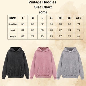 Op de afbeelding: Een maattabel voor vintage hoodies met afmetingen in centimeters voor maten S tot 4XL, inclusief schouder, borst en lengte. Onder de tabel staan drie hoodies met lange mouwen in zwart, oudroze en grijs met een gewassen afwerking.
