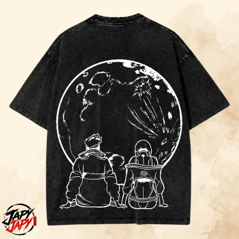 May include: A black acid-wash t-shirt featuring a large white circular graphic on the back. The design depicts two figures sitting and looking up at a detailed moon. The bottom left corner displays the text JAPY JAPY in a stylized black and red font.