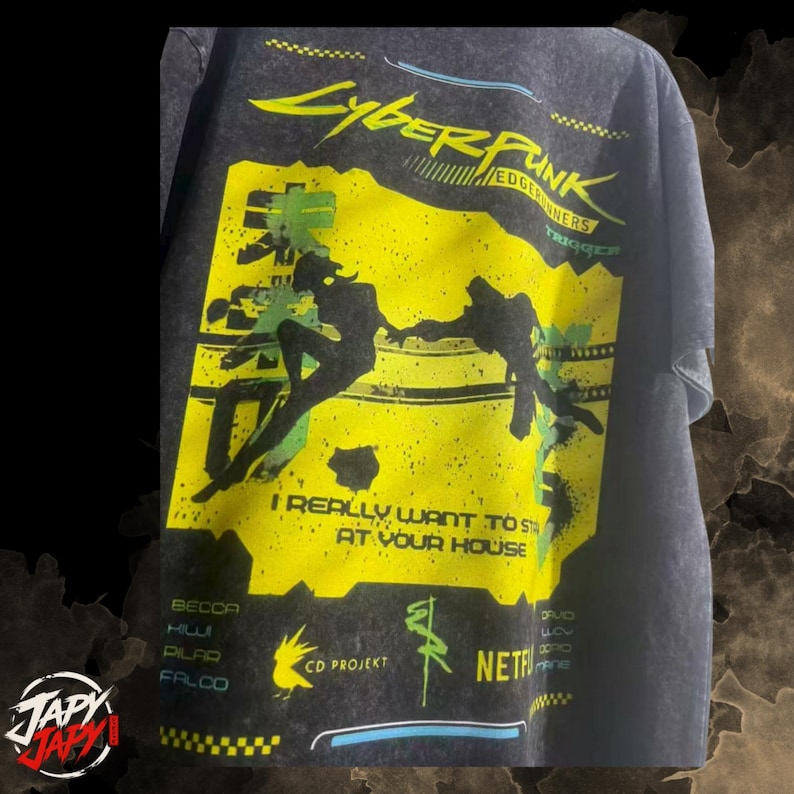 Op de afbeelding: Een antracietgrijs t-shirt met een grote gele grafische print. Het ontwerp bevat de tekst Cyberpunk Edgerunners, I Really Want To Stay At Your House en karakternamen. Het toont silhouetten van twee figuren die naar elkaar toe reiken.