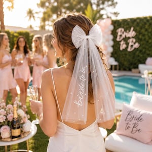May include: A white tulle hair bow veil featuring scattered faux pearls and the text "bride to be" in white script lettering. The accessory is attached to a hair clip and is designed to be worn at the back of the head for a celebratory event.