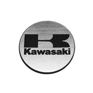 May include: A circular metal emblem with a brushed silver finish featuring the black Kawasaki logo and text centered on the surface. The design includes a stylized letter K graphic positioned above the word Kawasaki in a bold black font.