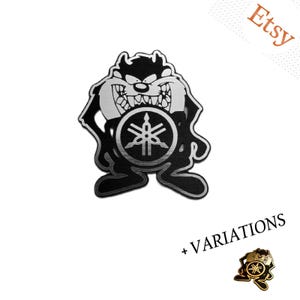 May include: A black and silver metal emblem featuring the Tasmanian Devil character with a prominent Yamaha logo centered on the chest. The design includes sharp teeth and a fierce expression. A smaller gold-toned variation is shown in the corner.