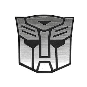 May include: A metallic silver emblem featuring the Autobot insignia from the Transformers franchise. The design consists of a stylized robot face with sharp, angular lines and a brushed metal texture, outlined in a bold, solid black border.